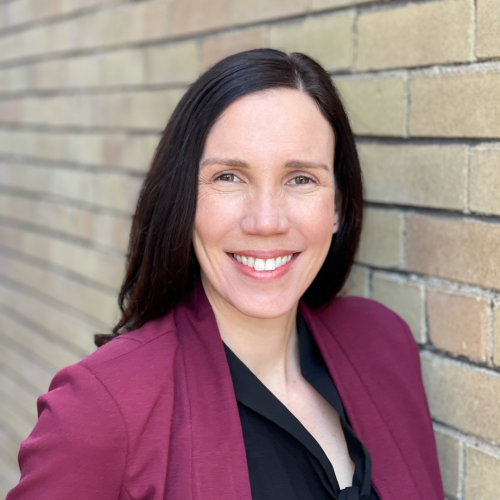 Photo of Professor Jennifer Campbell standing against a brick wall in a maroon blazer and black shirt.