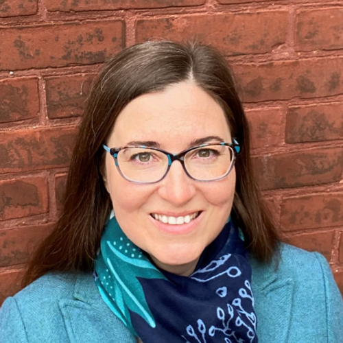 Photo of Alexandra Bolintineanu against a red brick wall wearing a navy scarf and teal blazer